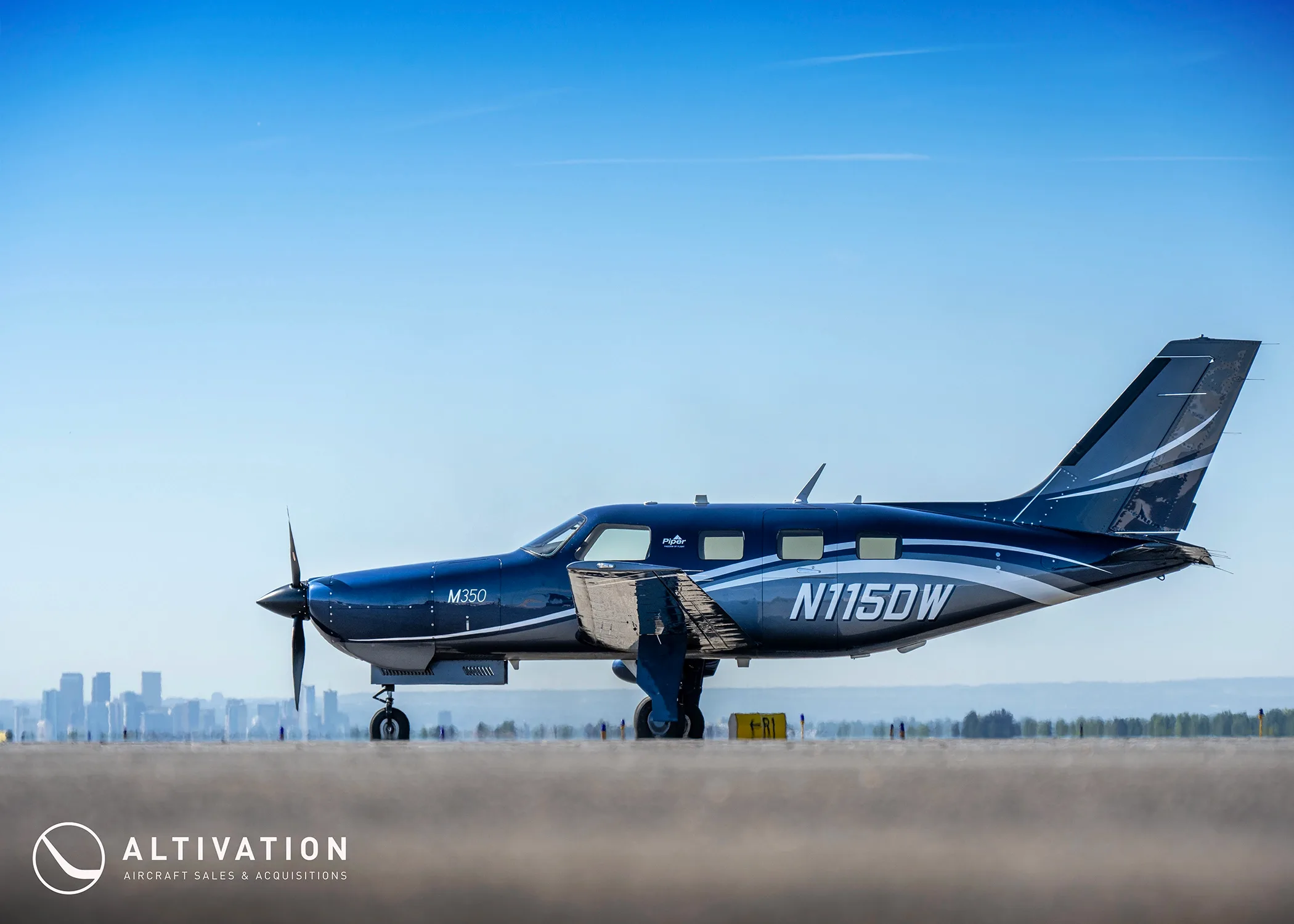 A dark blue Piper M350, tail number N115DW, parked on a tarmac with a city skyline in the background.