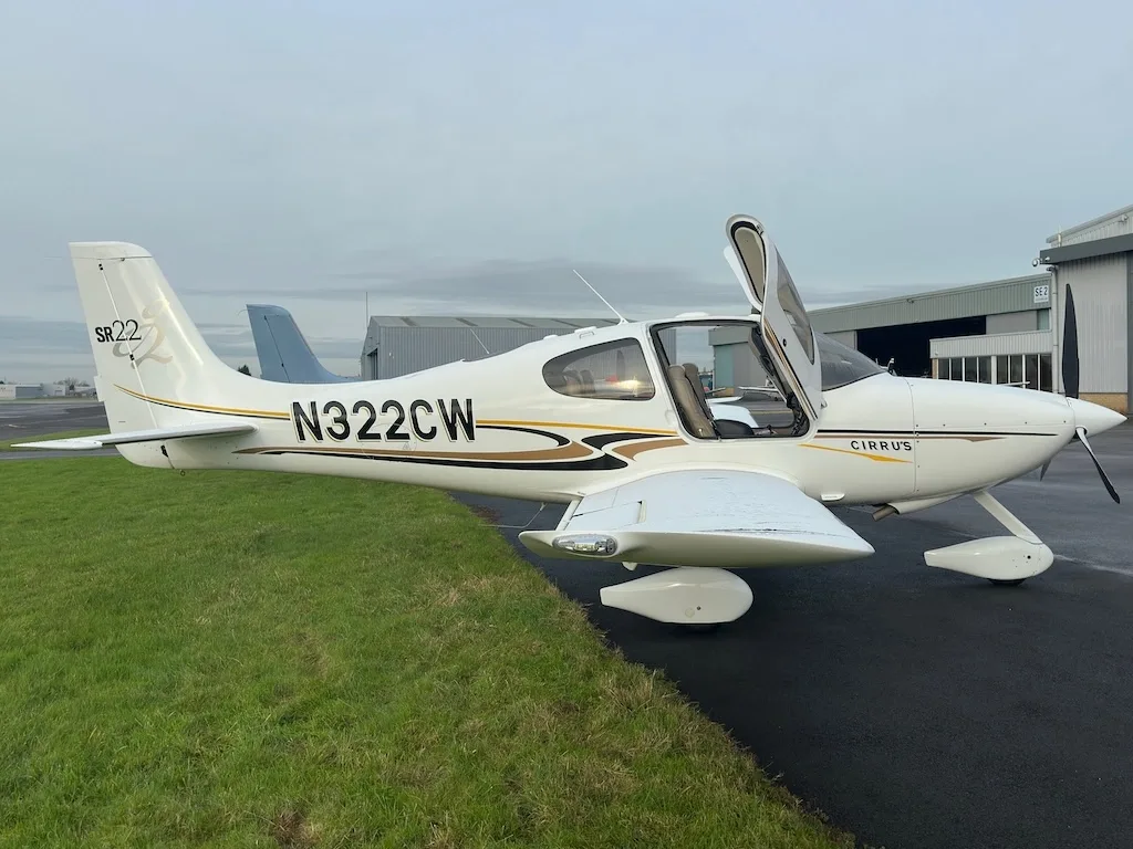 A side view of a white Cirrus SR22 aircraft with N322CW registration and open gull-wing doors, parked on a tarmac.