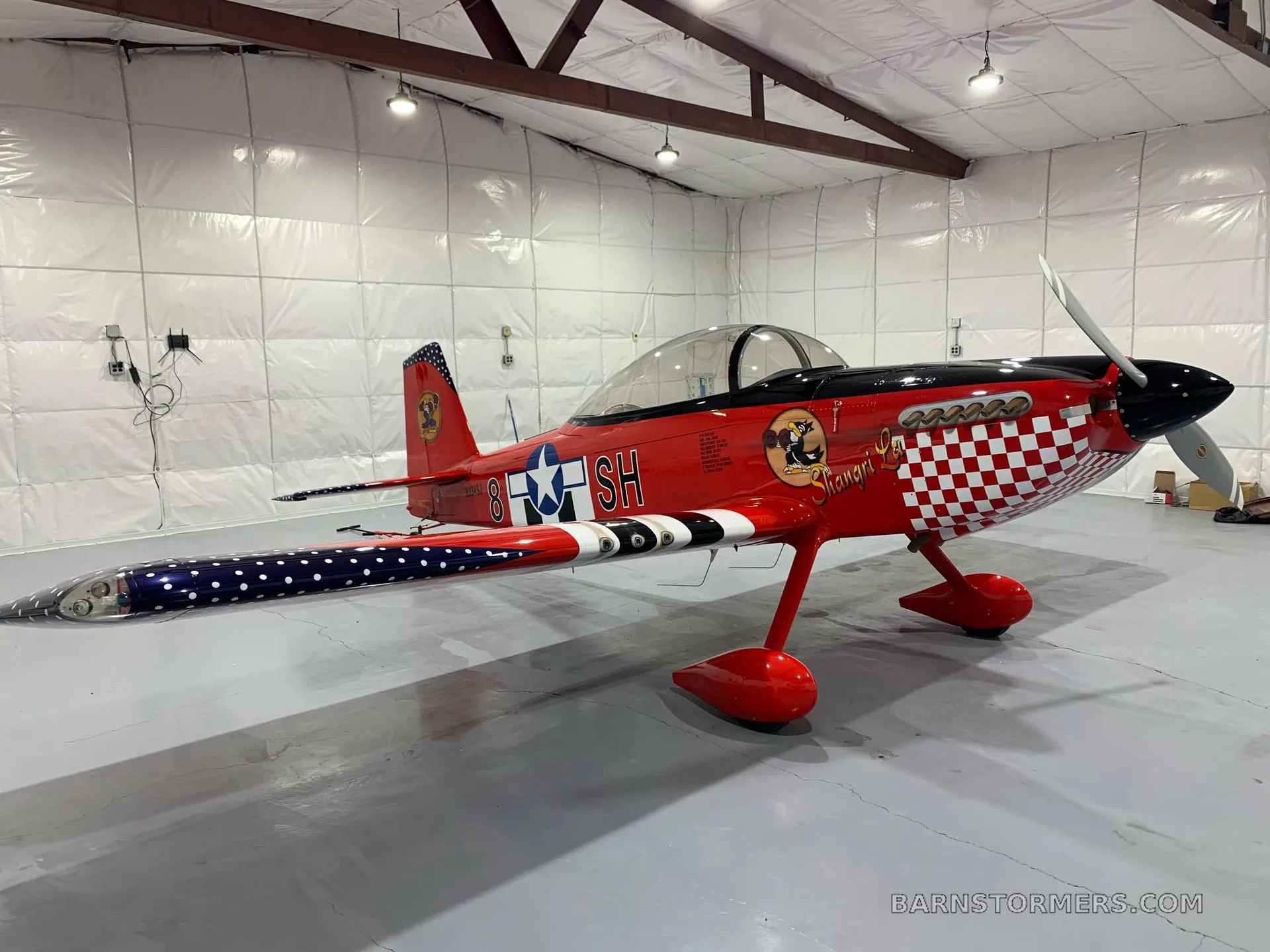 A red single-engine aircraft with a checkered nose and patriotic tail and wing tips, parked in a hangar.