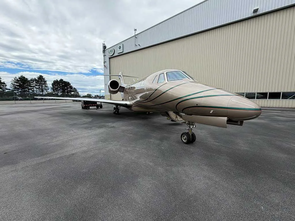 A front-quarter view of a beige business jet with green stripes parked on an asphalt apron next to a white hangar.