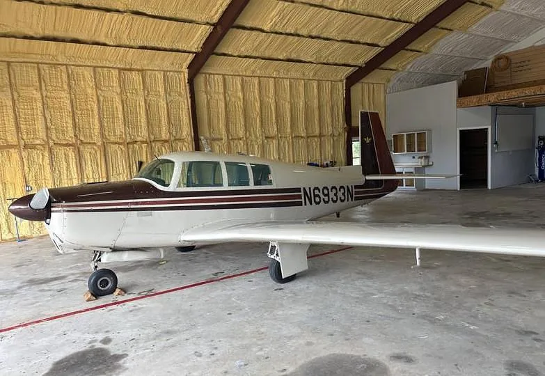 A white and brown Mooney M20 aircraft, registration N6933N, parked inside a hangar.