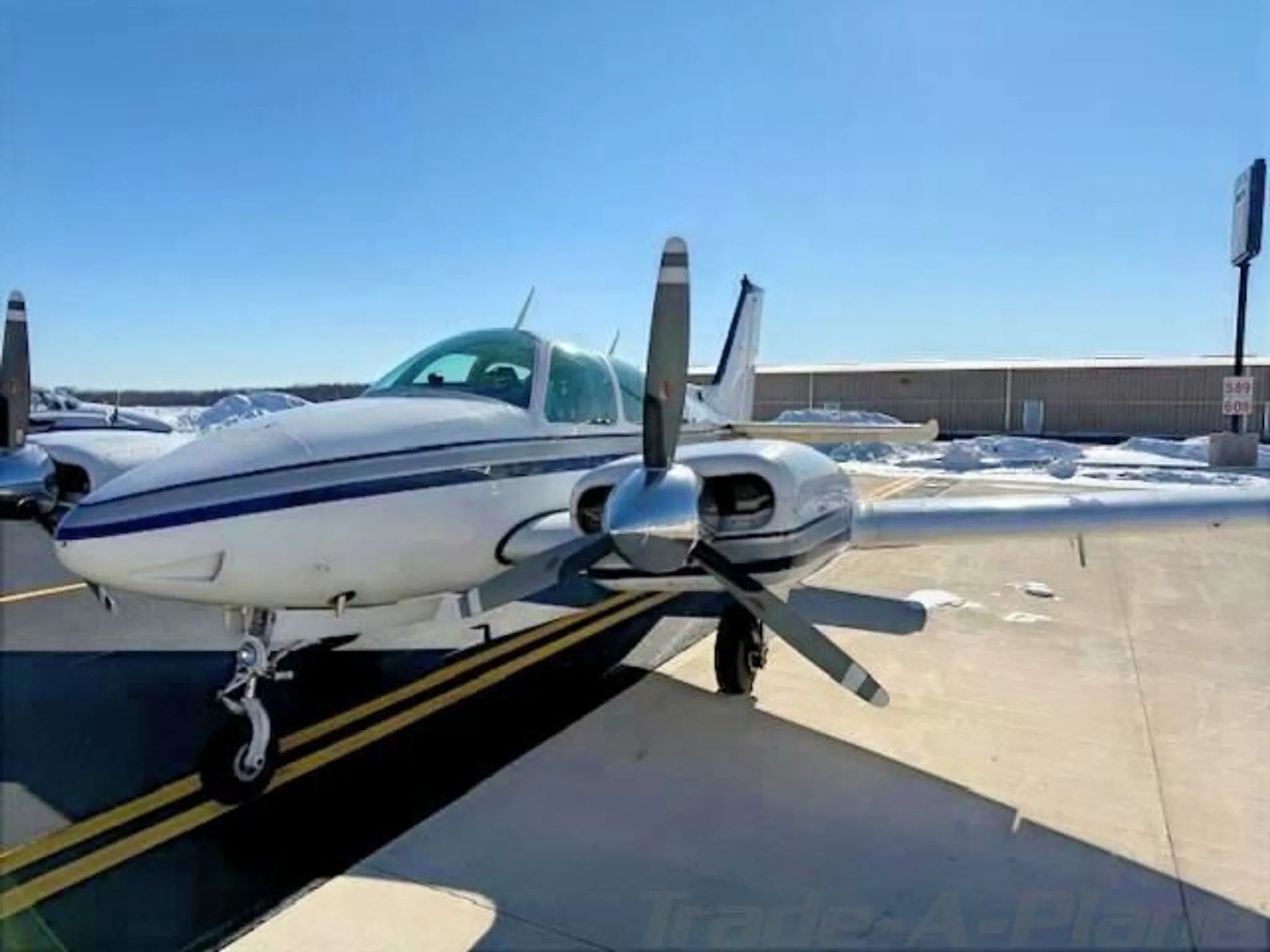 Front-right view of a white twin-engine aircraft with blue and grey stripes on a snowy tarmac.