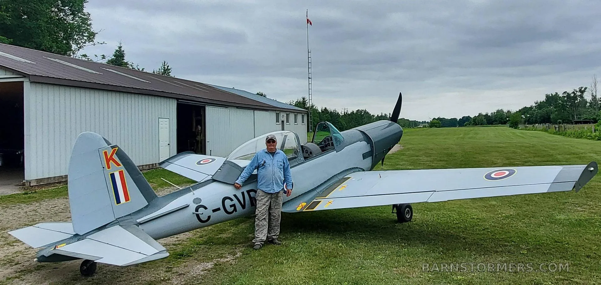 A light grey single-engine aircraft with Canadian registration C-GVDK and a person standing next to it on a grassy field.