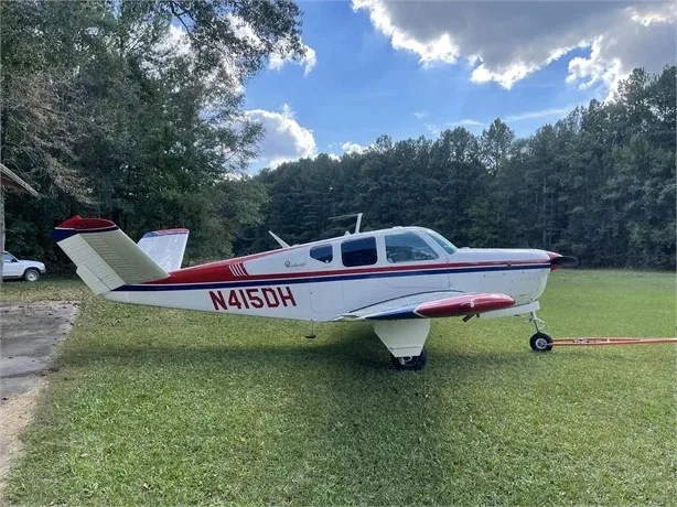 A side view of a white and red Beechcraft Bonanza N415DH parked on grass with trees in the background.