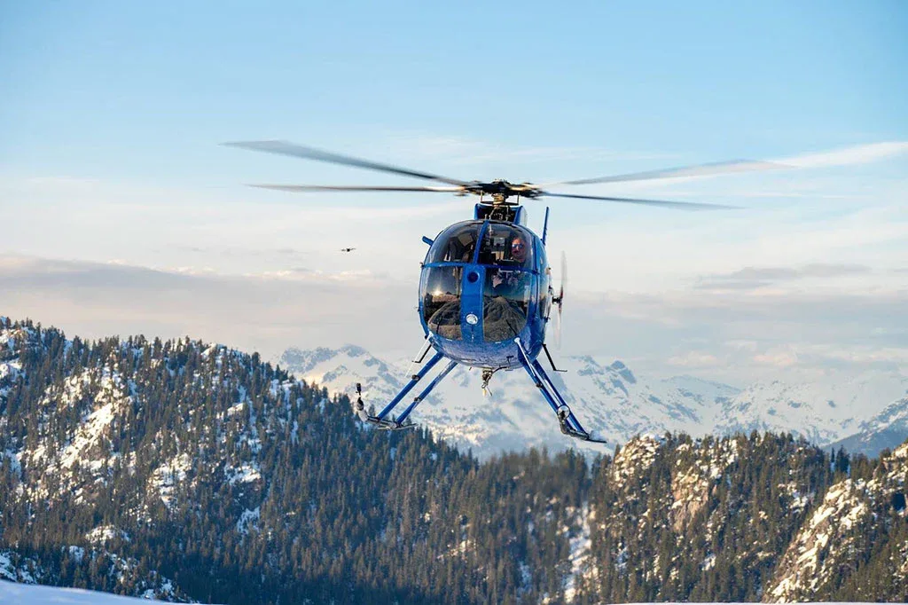 A blue helicopter with registration C-FJJ0 flies over snow-dusted, forested mountains under a clear sky.