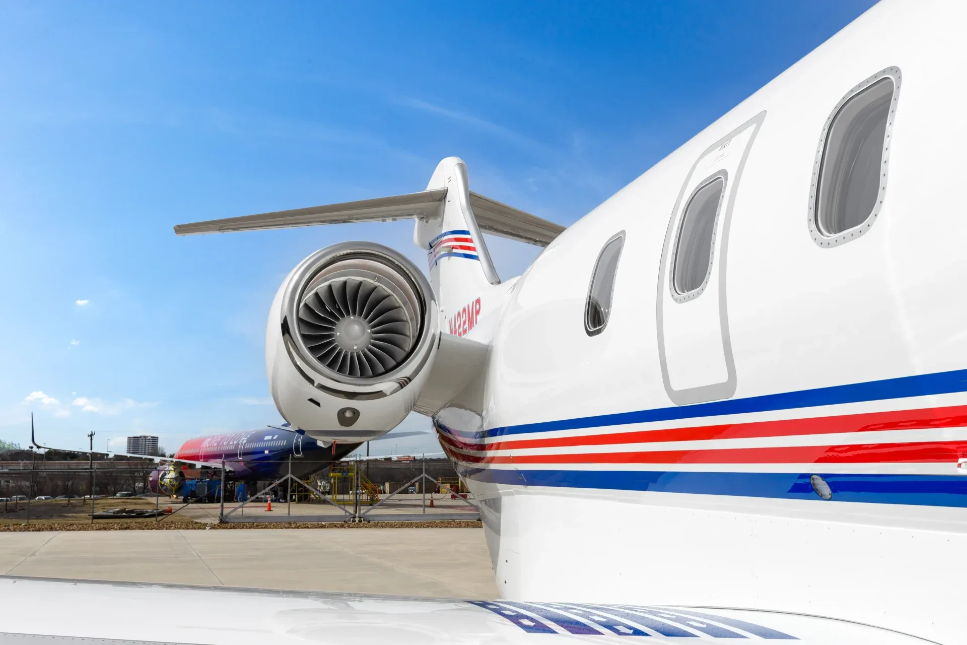 A close-up view of the rear fuselage and engine of a white business jet with red and blue stripes, parked on an airfield.