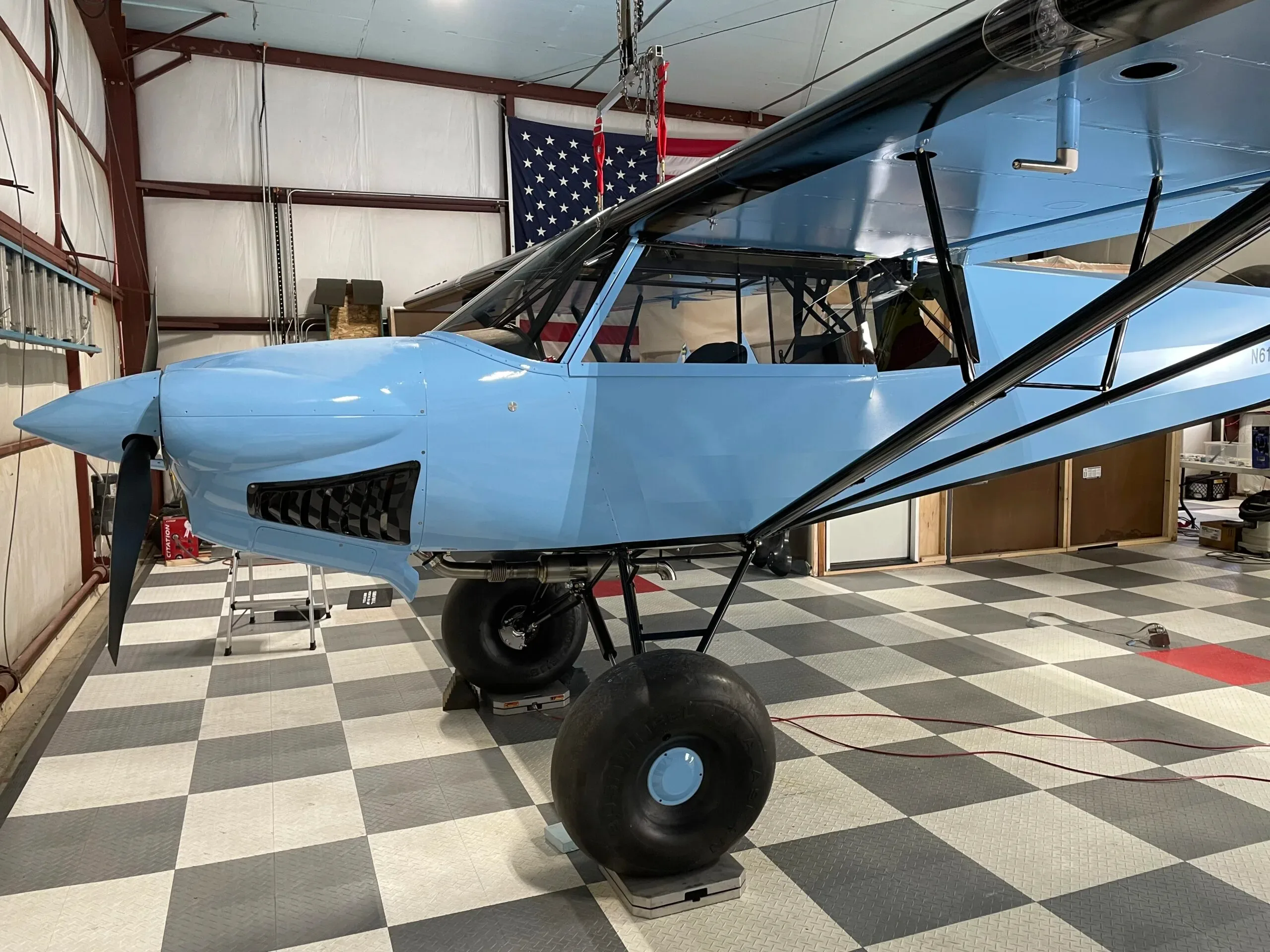 A side view of the light blue "Bluebird" aircraft with large tundra tires, parked on a checkered floor in a hangar.