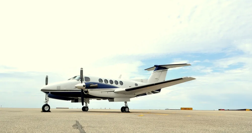 A white and blue twin-engine turboprop aircraft, likely a Beechcraft King Air, parked on an airport tarmac under a partly cloudy sky.
