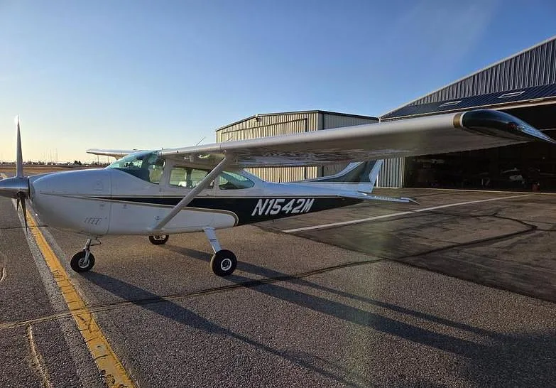 A white and green Cessna aircraft (N1542M) is parked on an asphalt runway with hangars in the background.