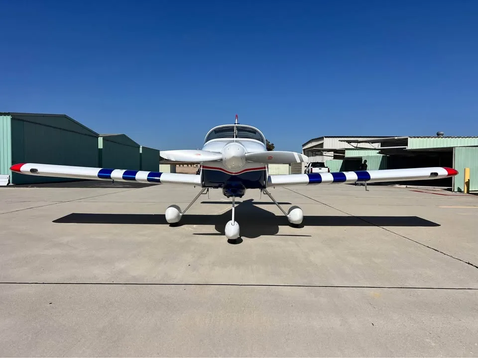 A white, blue, and red single-engine aircraft with striped wings and tail, viewed from the front on an airfield.