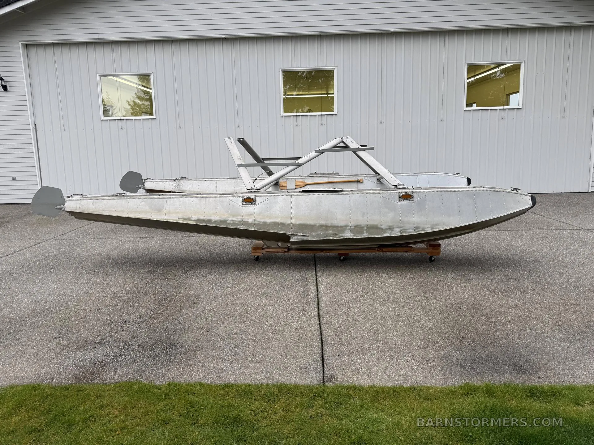 A pair of aluminum aircraft floats with mounting struts, seen from the side, on a concrete driveway in front of a white building.