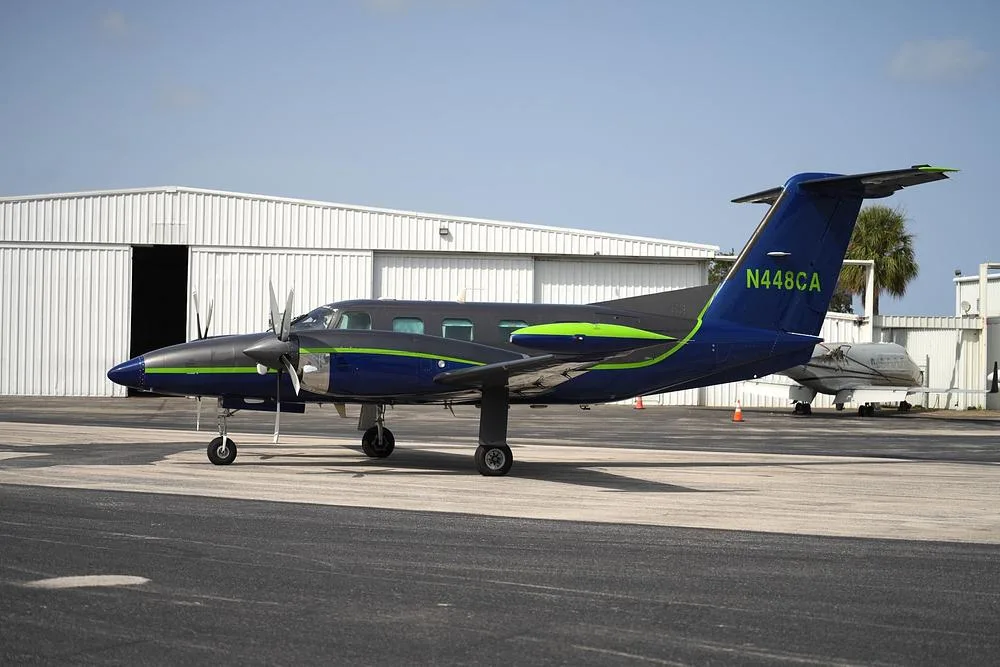 A side view of a dark blue and grey twin-engine turboprop aircraft with a green stripe, tail number N448CA, parked on an airfield.