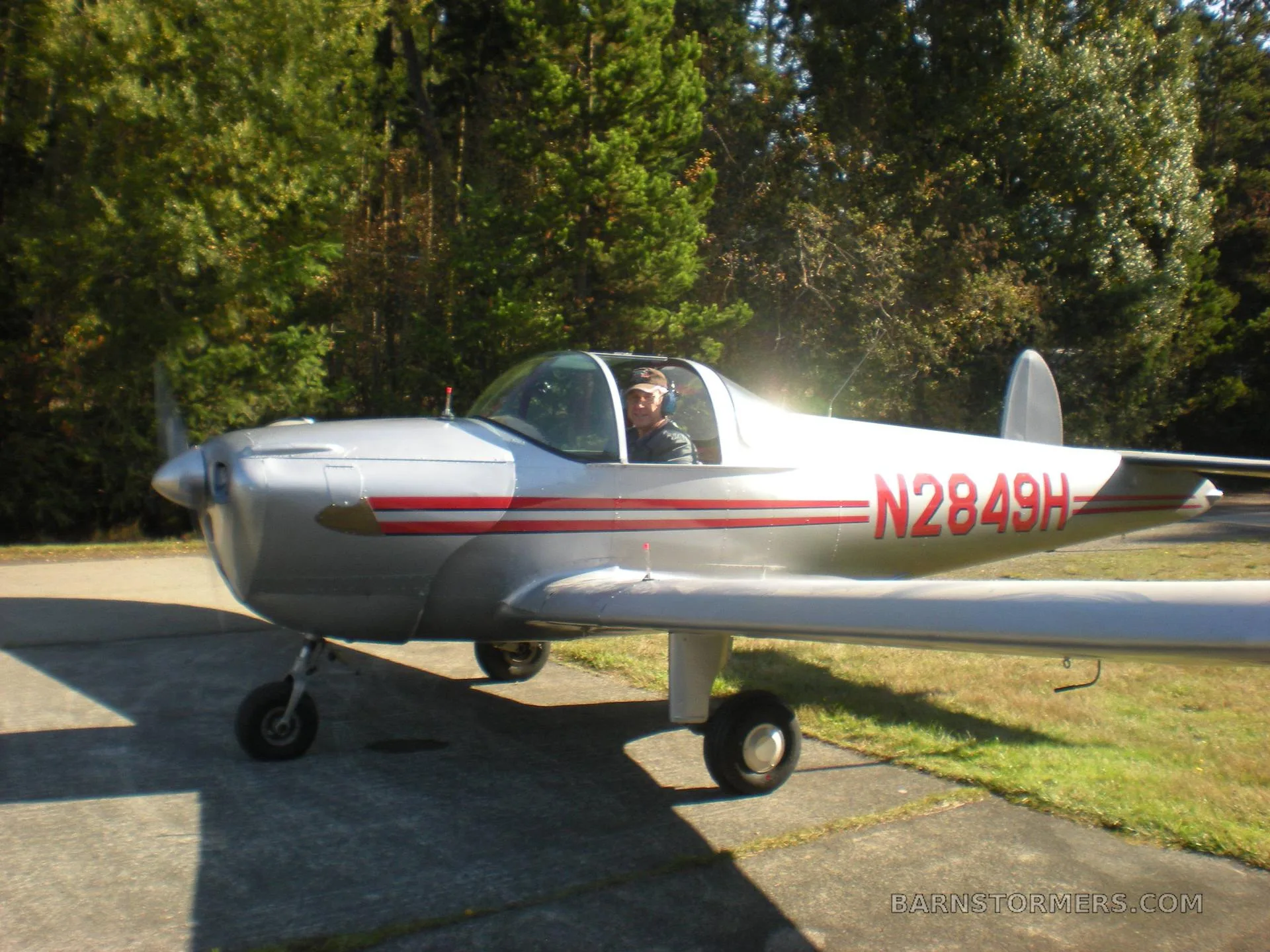 A silver Ercoupe aircraft with red stripes and tail number N2849H, with a pilot in the cockpit, on a paved surface surrounded by trees.