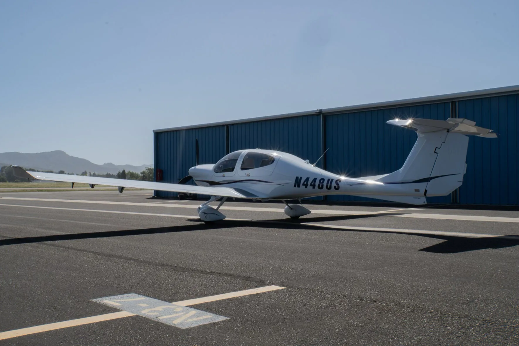 A white Diamond DA40 Star XL aircraft with purple and grey stripes, registration N448US, parked on an tarmac in front of a blue hangar.