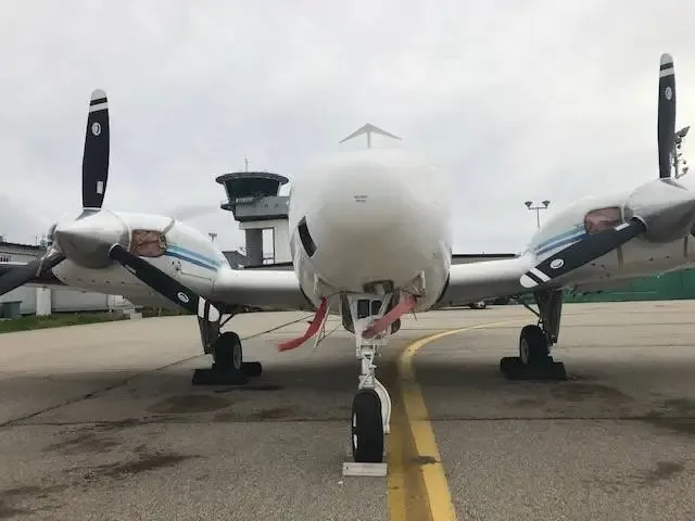 Head-on view of a white twin-engine propeller aircraft with red pitot tube covers on an airport tarmac.
