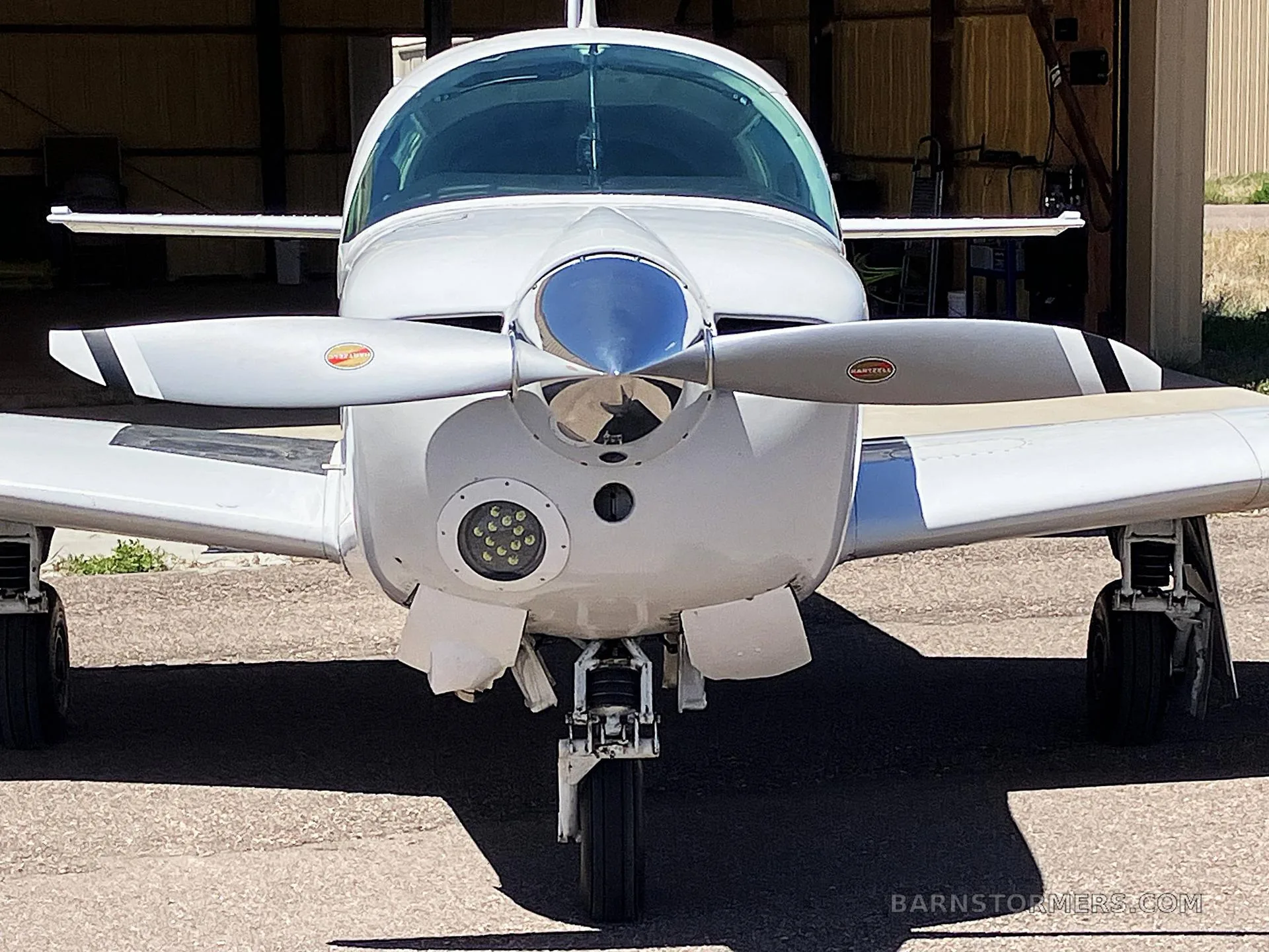 A head-on view of a white Mooney M20J aircraft with a three-bladed propeller, parked in a shaded area.