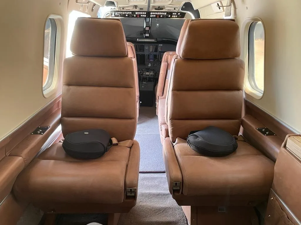 The interior cabin of an aircraft, showing two rows of brown leather seats facing forward towards the cockpit.