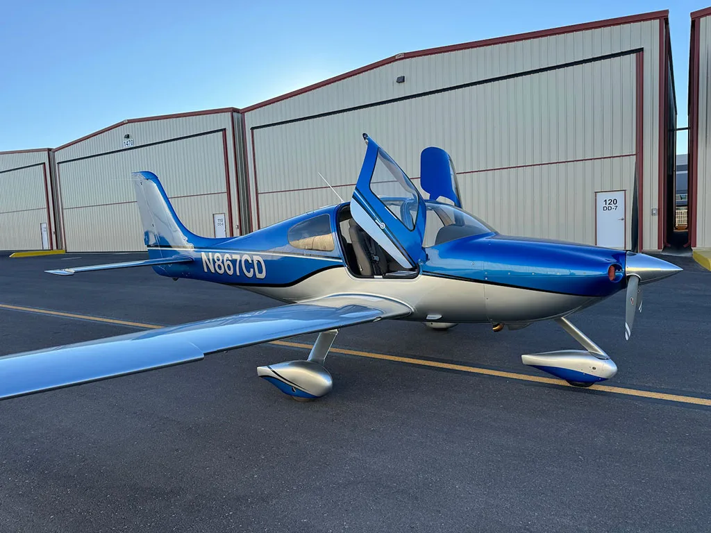 A blue and silver Cirrus SR22 aircraft with registration N867CD parked on an airfield with its doors open.