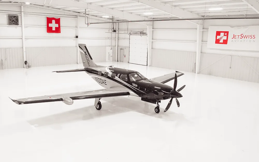 An aerial view of the black and silver Piper M600/SLS aircraft (N600AE) parked centrally in a spacious, white hangar.