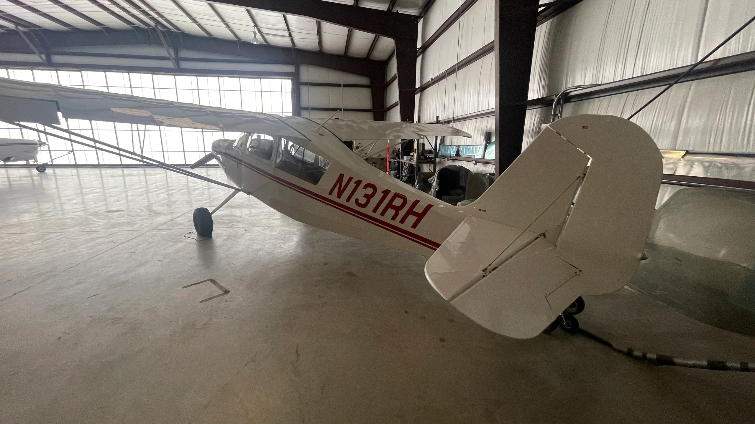 The rear-right view of a white taildragger aircraft with red stripes and registration N131RH, parked inside a hangar.