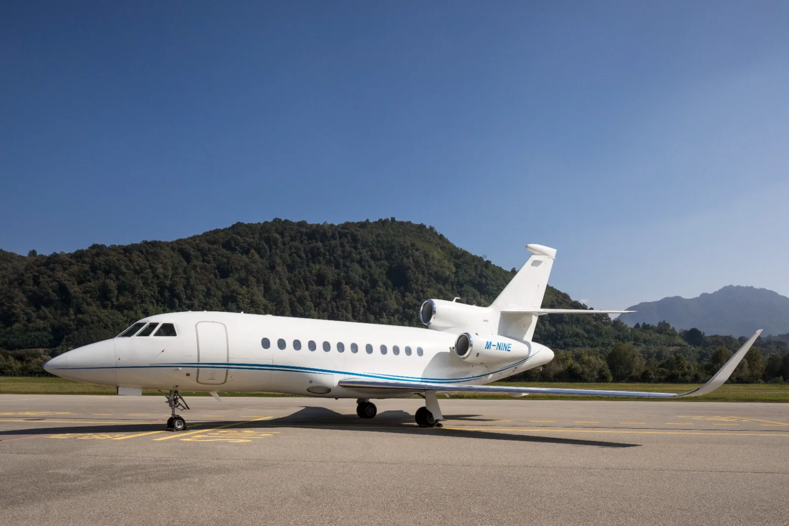 A white Dassault Falcon business jet, registration M-NINE, parked on an airfield with mountains in the background.