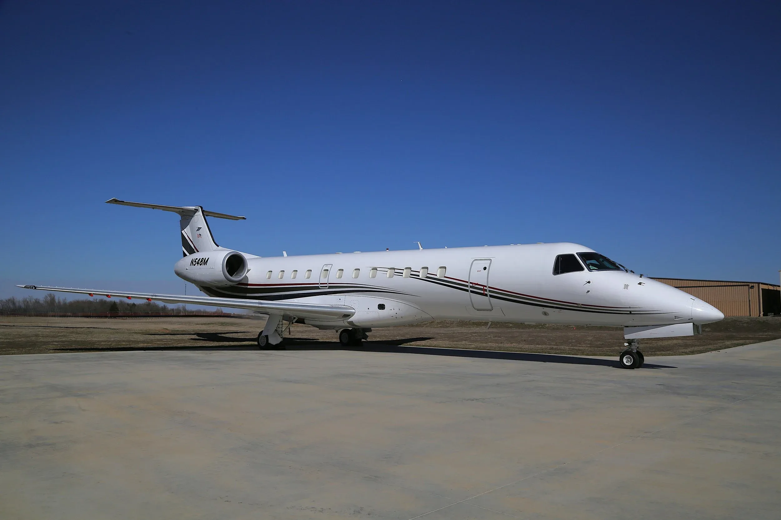 A white Embraer Legacy business jet with black, red, and gold stripes is parked on a tarmac under a clear blue sky.