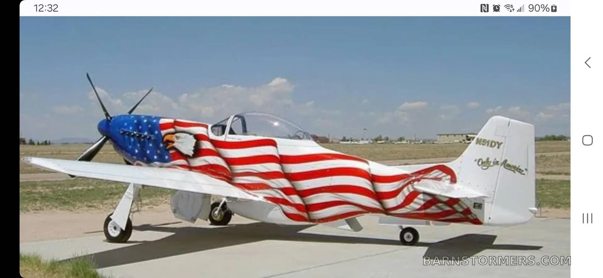 An American flag-painted P-51 Mustang replica parked on an airfield under a partly cloudy sky.