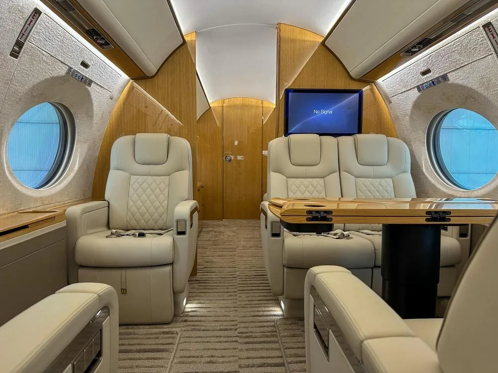 An aircraft cabin interior with light leather seats, a wooden table deployed between two seats, and a monitor displaying "No Signal".