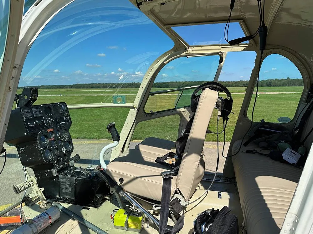 The cockpit of the Hiller UH-12 helicopter showing the instrument panel and controls with a view of the airfield.