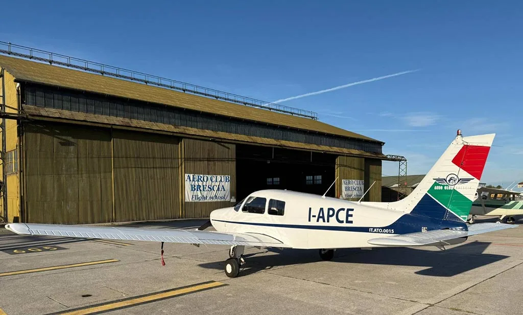 A white single-engine propeller aircraft with "I-APCE" registration parked in front of a large green hangar with "Aero Club Brescia Flight School" signs.