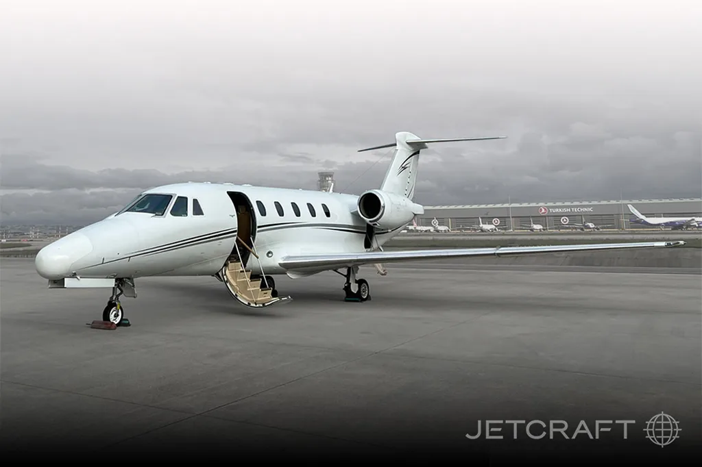 A white Cessna Citation business jet with its door open and stairs deployed, parked on an airfield under a cloudy sky.