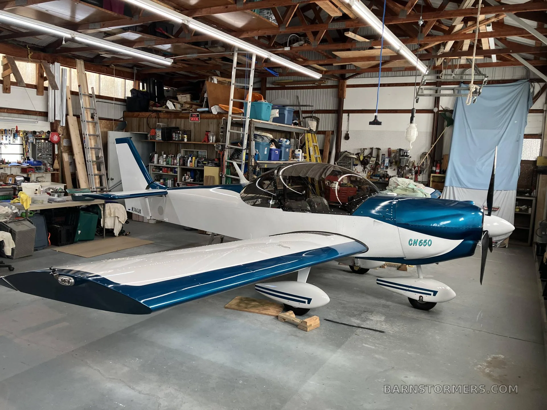 A white and blue Zenith CH650 aircraft with registration N744JK is parked inside a cluttered workshop.