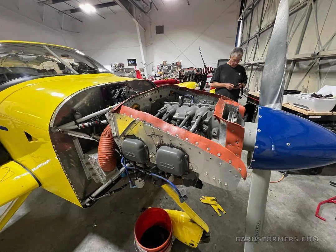 A yellow aircraft with a blue propeller tip has its engine cowling open, revealing the engine, while a person works nearby in a hangar.