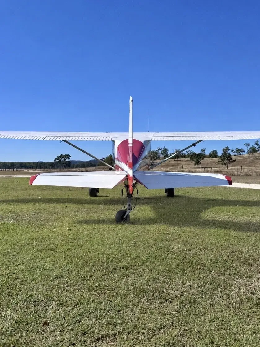 A rear view of a red and white single-engine propeller aircraft parked on a grassy field with distant trees.