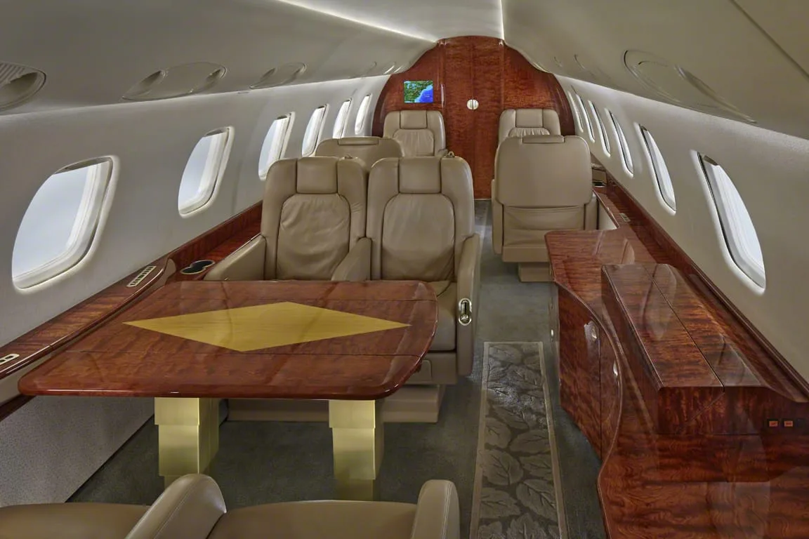 The interior of a private jet cabin featuring beige leather seats, a wooden table, and wood paneling.