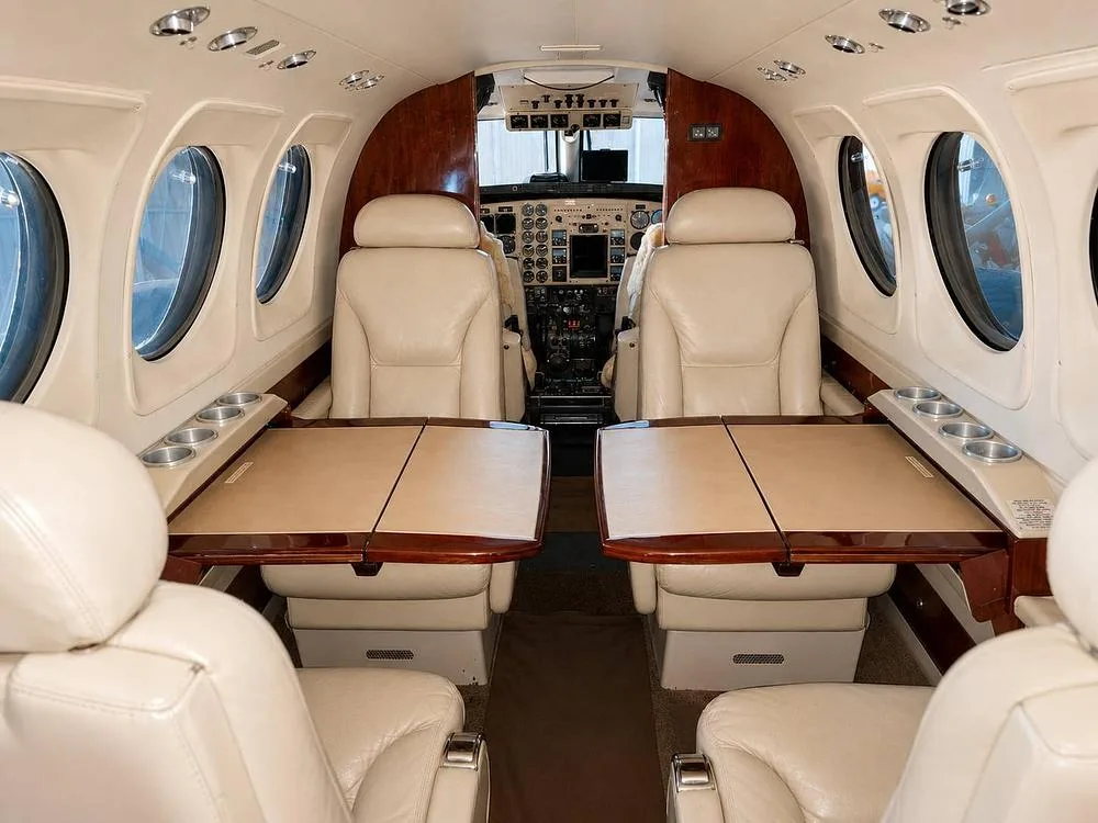 The aircraft cabin features beige leather seats, two fold-out tables, and wood paneling, looking towards the cockpit.