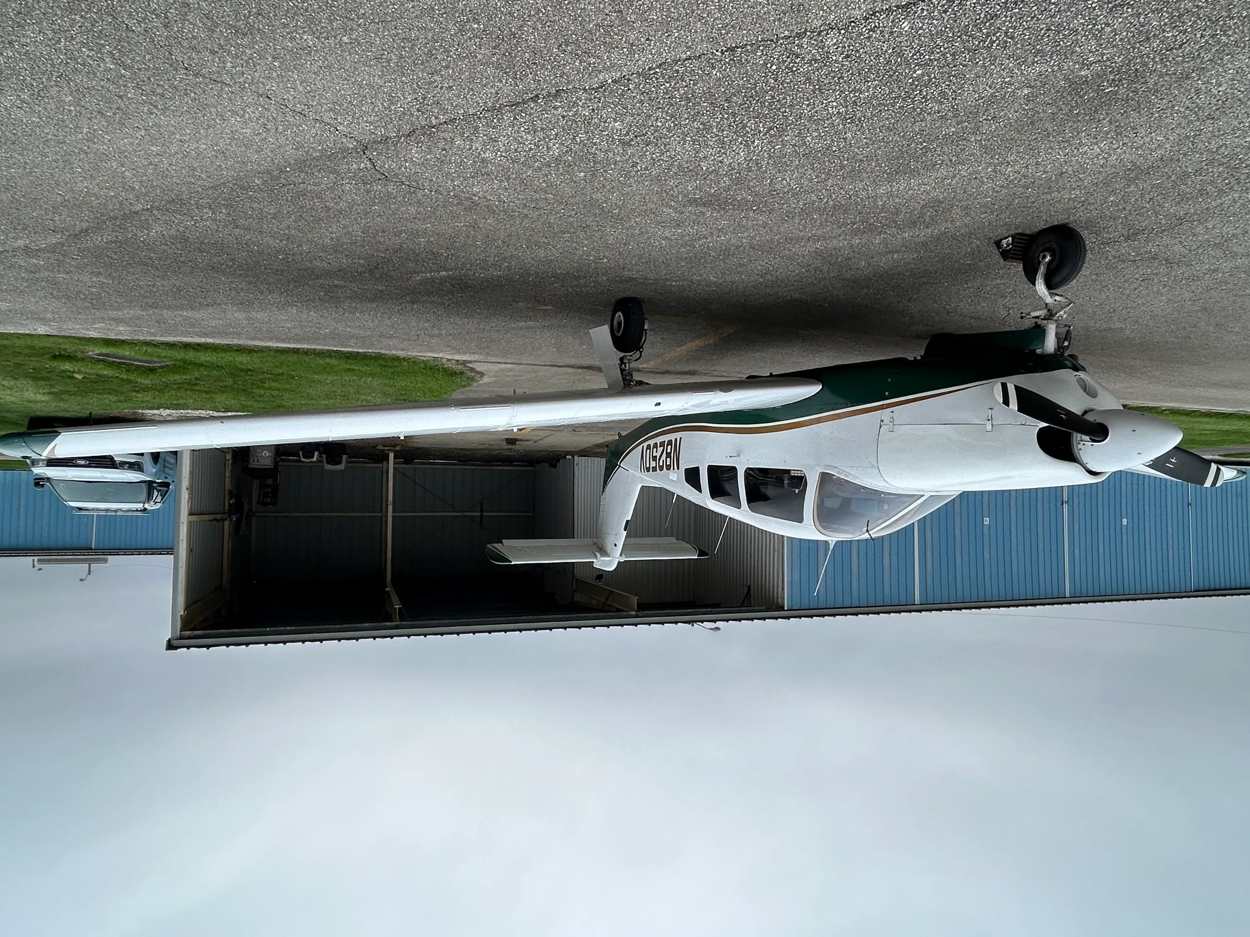 A white and green single-engine aircraft, N8250V, parked on an asphalt ramp in front of a blue hangar.