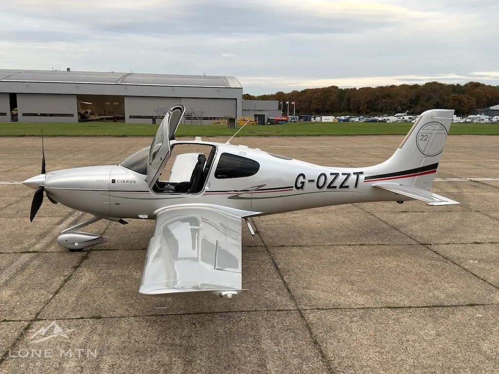 A silver Cirrus SR22T aircraft with registration G-OZZT parked on an airfield tarmac, viewed from the side.