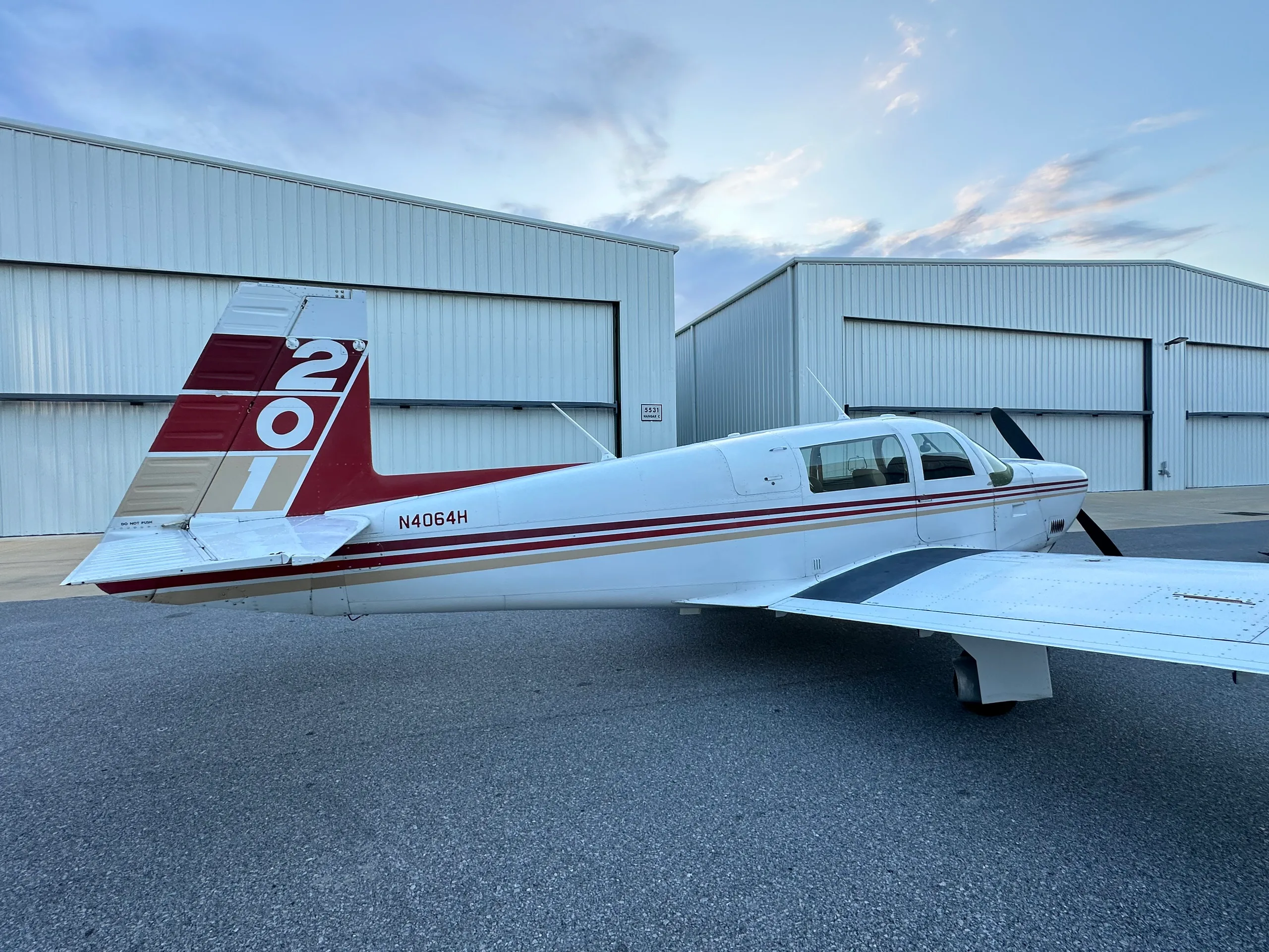 A rear-side view of a white single-engine aircraft with red and gold stripes on the tail and fuselage, parked outside hangars.