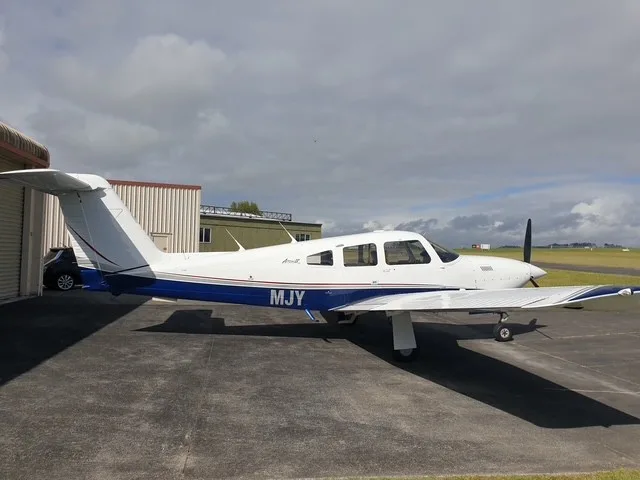 A white and blue Piper Arrow aircraft with registration MJY parked on a tarmac next to a hangar.
