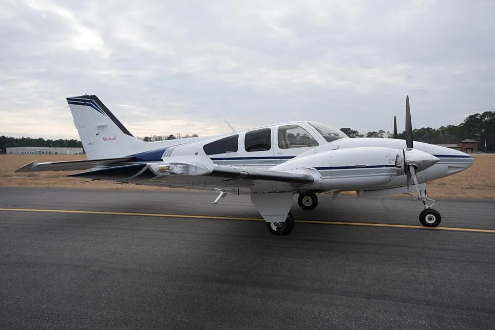 A side view of a white twin-engine propeller aircraft with blue and grey stripes parked on an asphalt runway.