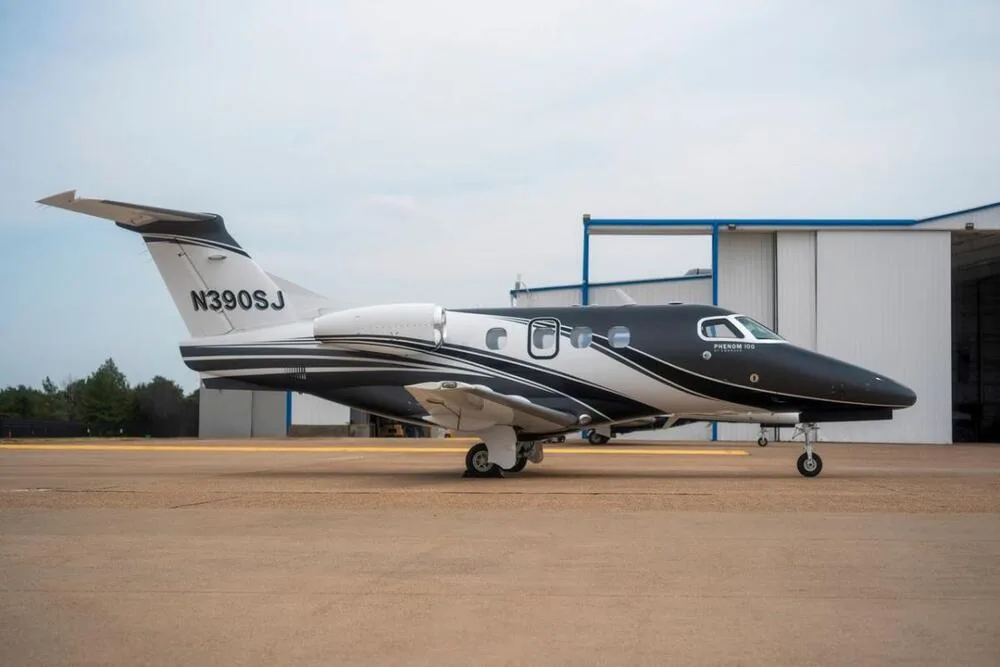 A side view of a black and white Embraer Phenom 100 business jet (N390SJ) parked on an airport tarmac next to a hangar.