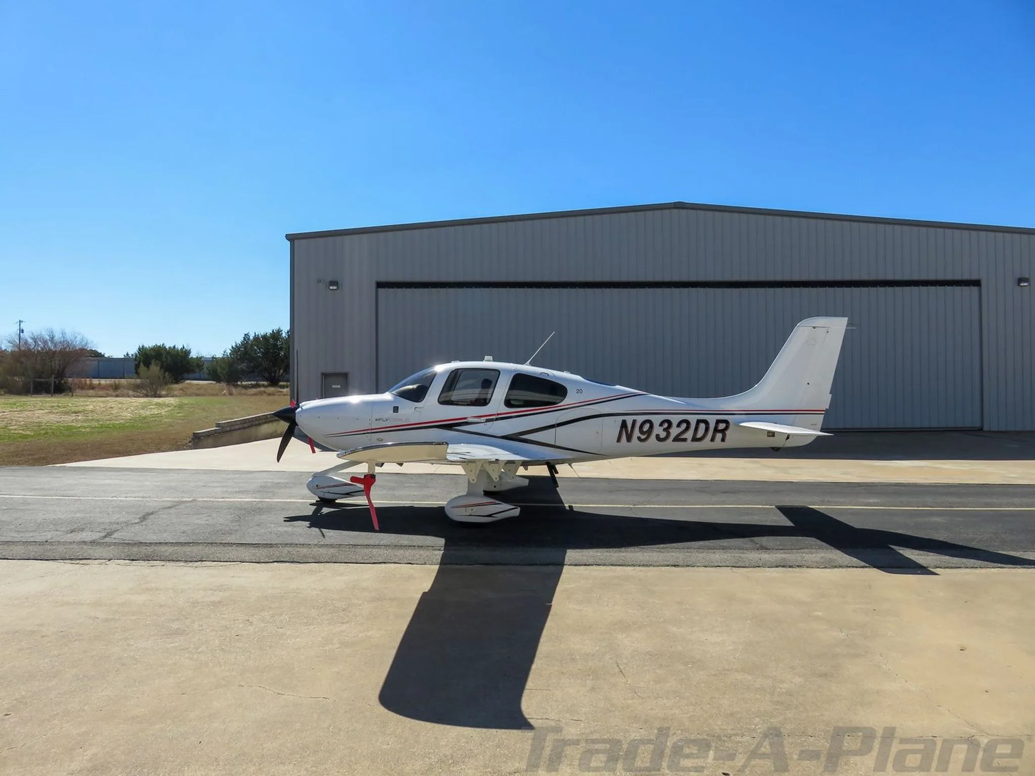 White single-engine aircraft with red and black stripes, registration N932DR, parked on a tarmac in front of a hangar.