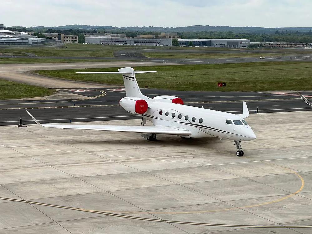 A white Gulfstream G650 business jet with red engine nacelles parked on a concrete tarmac near a runway.