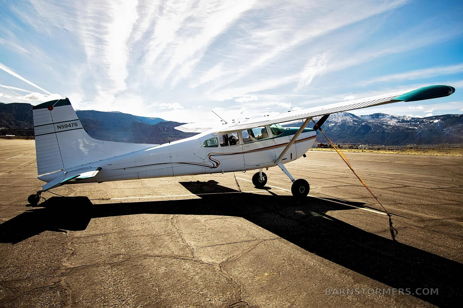 A white and teal single-engine propeller aircraft with registration N5947S parked on an airfield under a dramatic sky with mountains in the background.