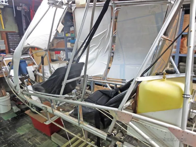 A partially assembled ultralight aircraft frame with two black seats and a yellow fuel tank in a workshop.