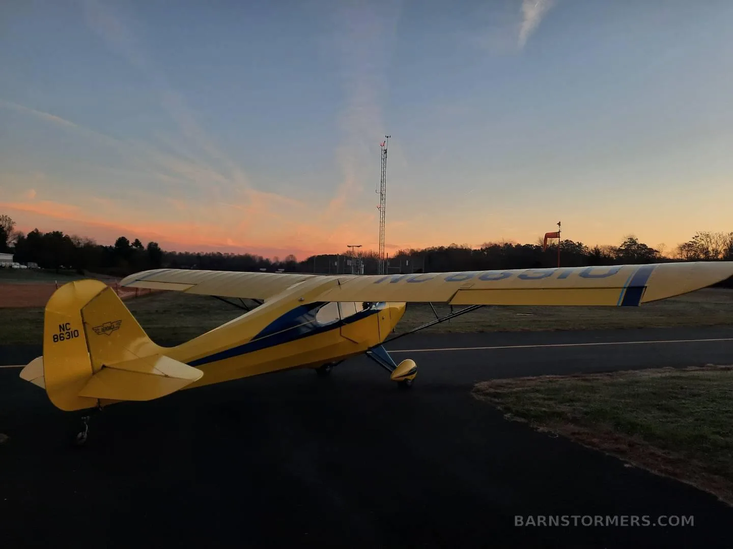 A yellow and blue Aeronca Champ aircraft is seen from the rear, parked on an asphalt ramp at sunset.