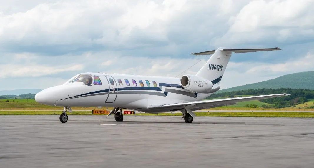 A white Cessna Citation CJ3 business jet (N906MC) with blue and grey stripes is parked on a tarmac under a cloudy sky.