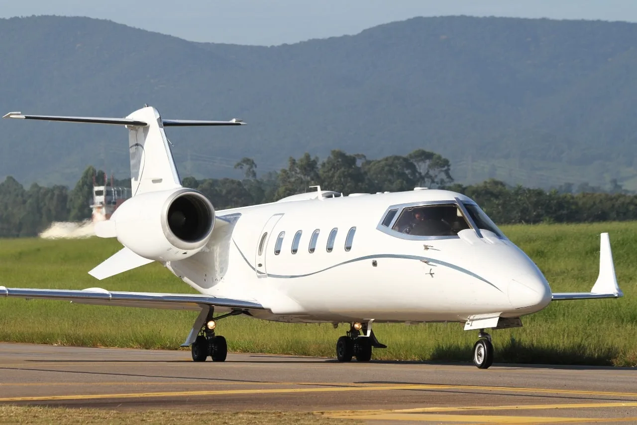 A white business jet with blue pinstripes is seen from a front-side angle on a runway with green grass and hills.