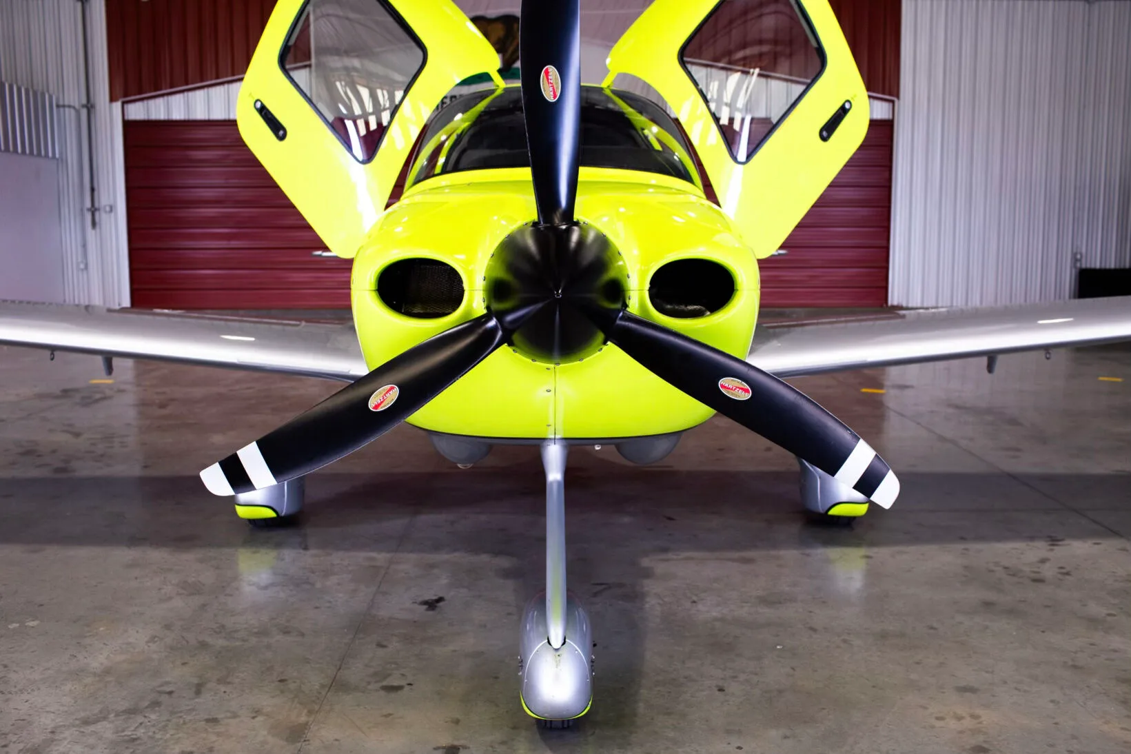 Front view of a bright yellow and silver Cirrus SR20 aircraft with open doors in a hangar.